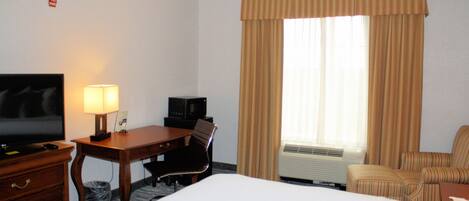 A hotel room with a bed, a desk with a lamp, a television, a chair, and a window with curtains.