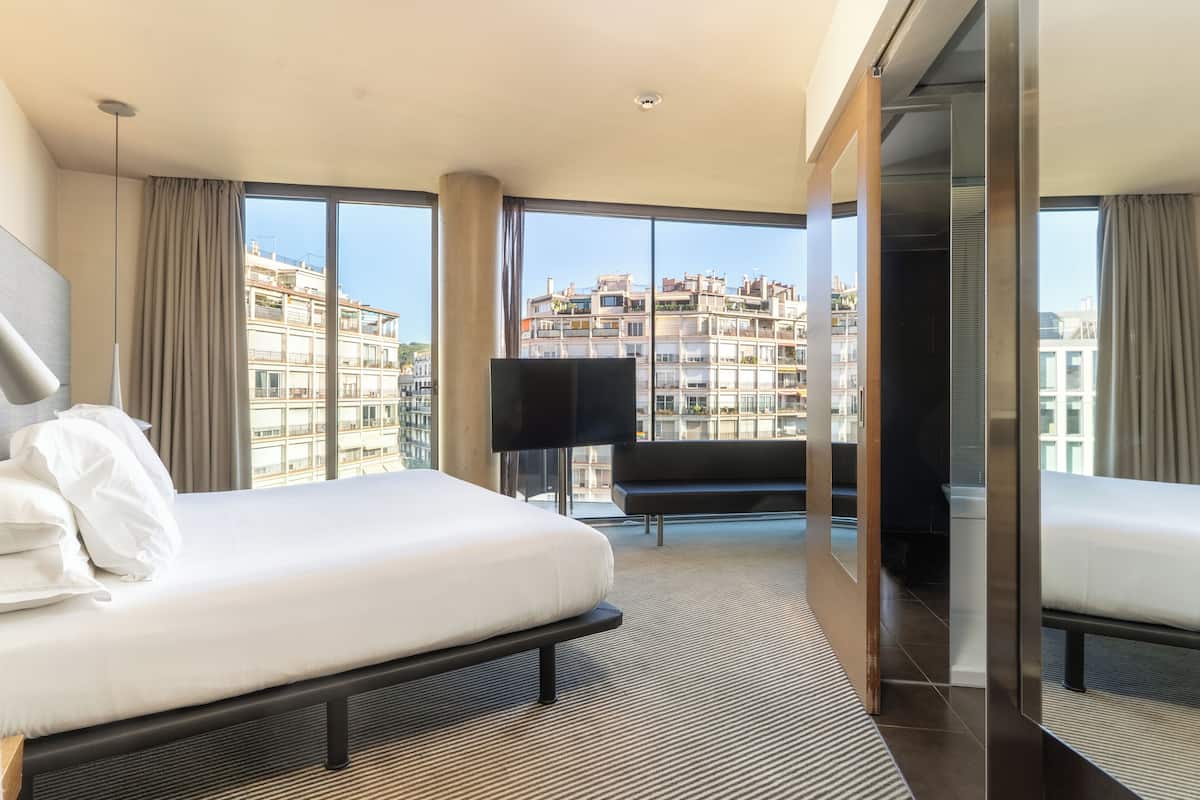 A modern hotel room with a large bed, a flat-screen TV, and a view of the cityscape through large windows.