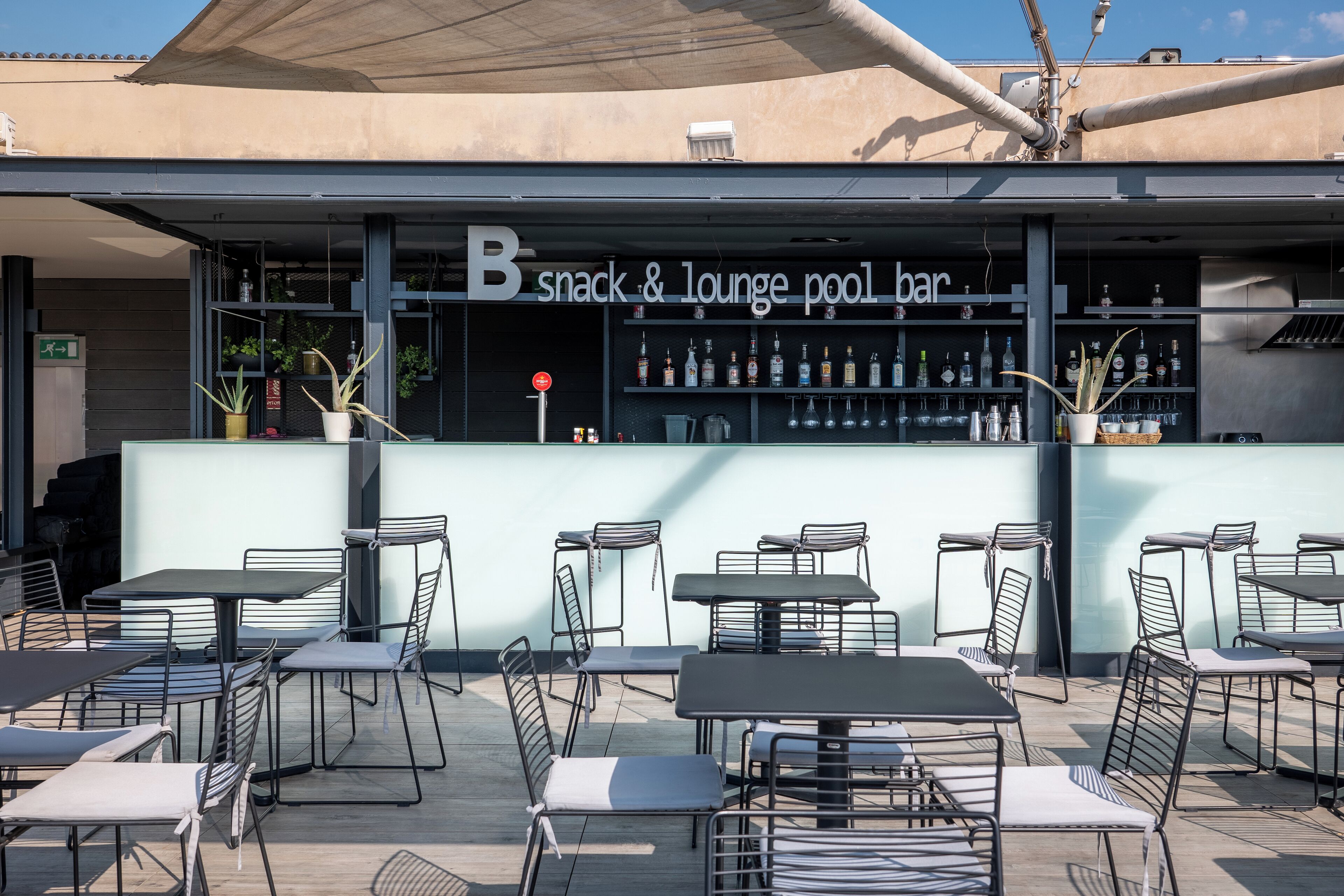 Un espace extérieur aménagé avec des tables et des chaises, un bar avec des étagères remplies de bouteilles, et une enseigne indiquant « B snack & lounge, bar au bord de la piscine ».