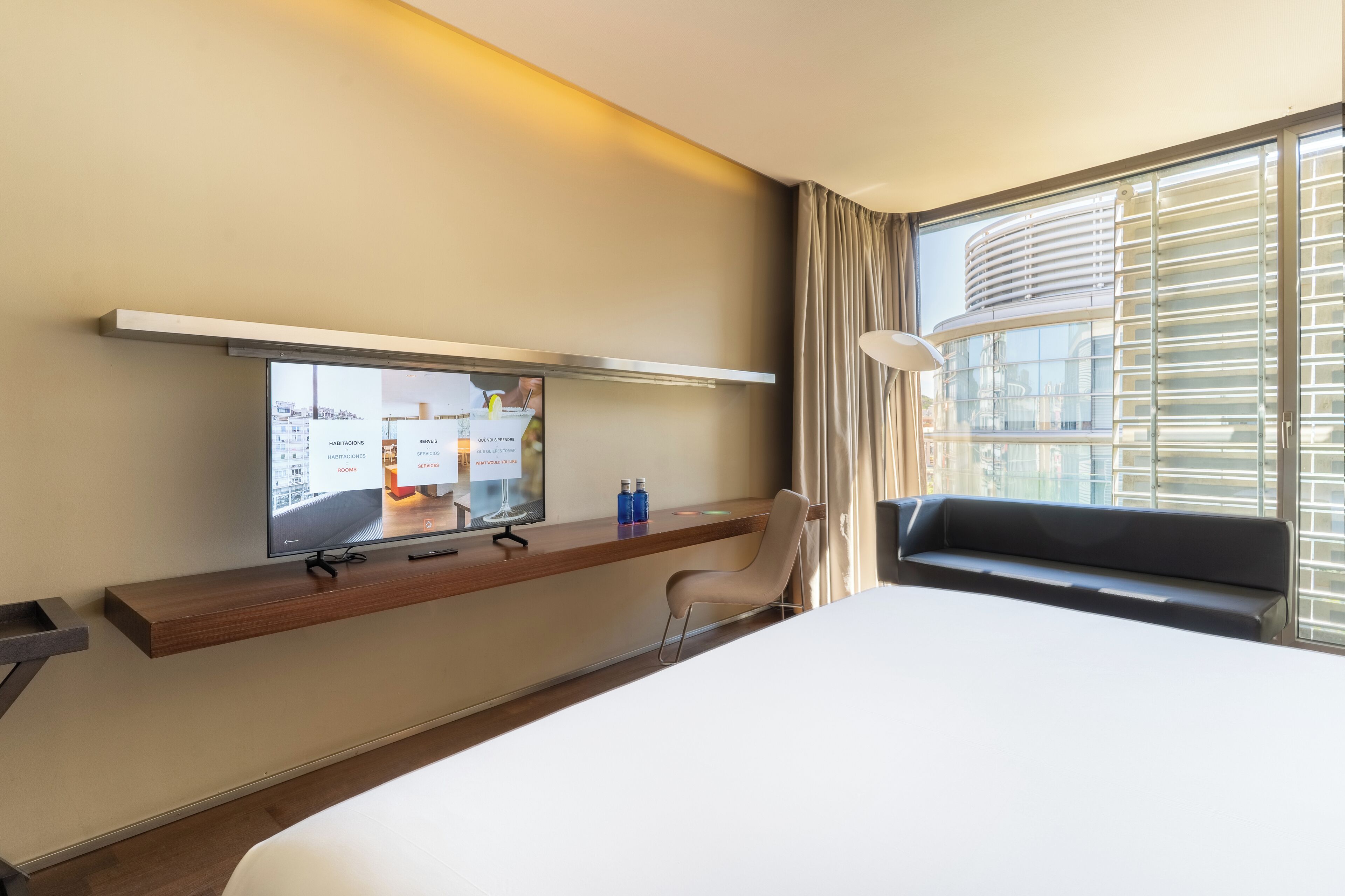 A hotel room with a bed, a TV mounted on a wooden shelf, a chair, and a window with blinds.