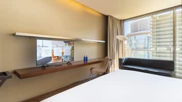 Habitació d'hotel amb un llit, una televisió muntada en una estant de fusta, una cadira i una finestra amb persianes.