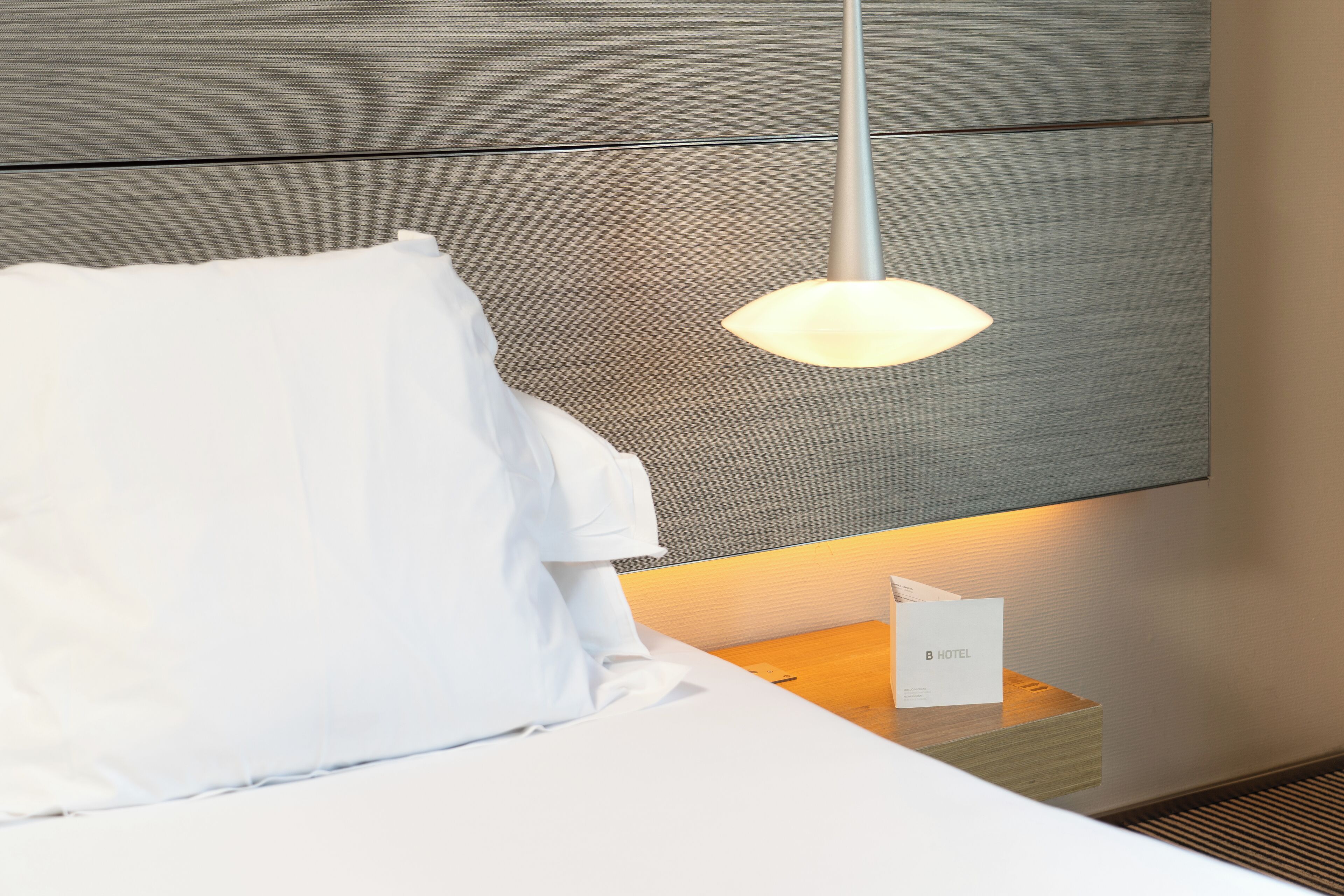 A neatly made bed with white linens, a wooden headboard, and a bedside table with a lamp and a hotel business card.