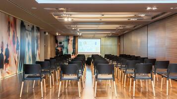 Uma sala de conferências com fileiras de cadeiras, uma tela de projetor e uma apresentação na parede.