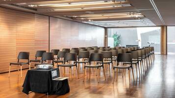 Sala de conferências com fileiras de cadeiras, um projetor sobre uma mesa e um grande janelão.