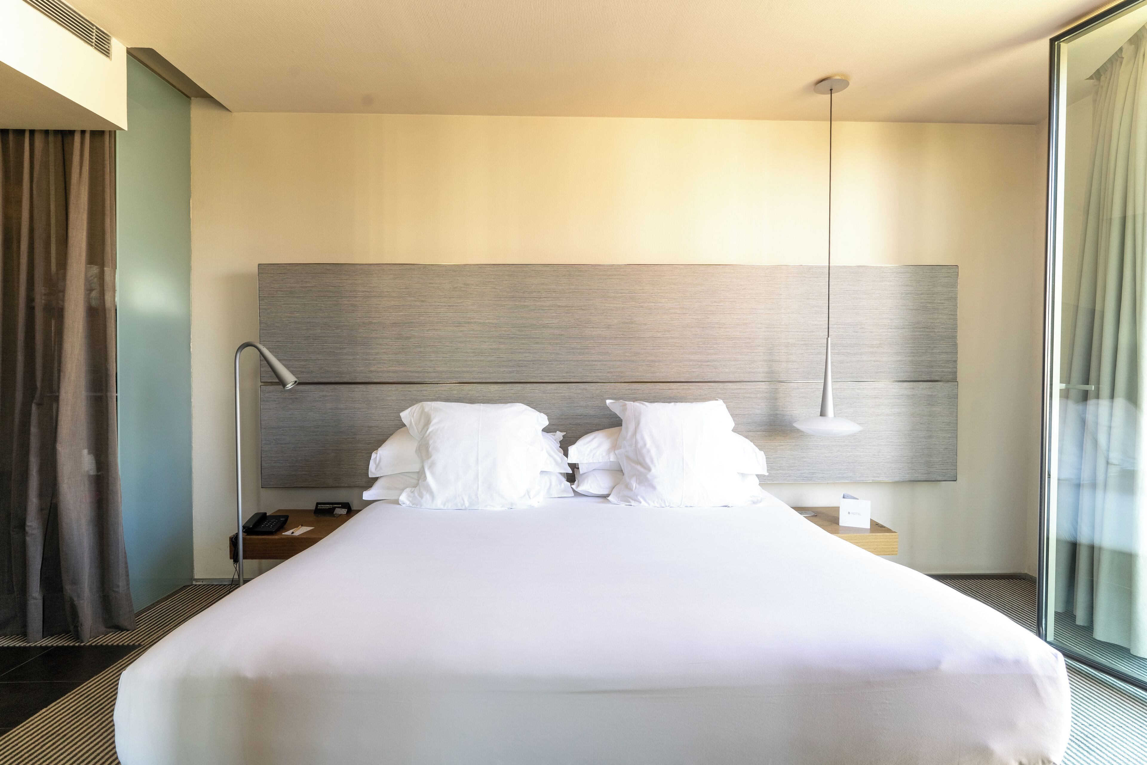 Een moderne hotelkamer met een groot bed, een nachtkastje met een lamp en uitzicht door een groot raam.