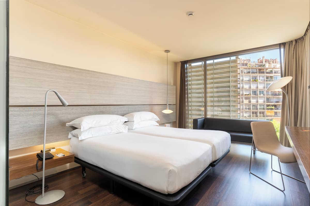 A hotel room with two beds, a desk, a chair, a lamp, and a view of the city.