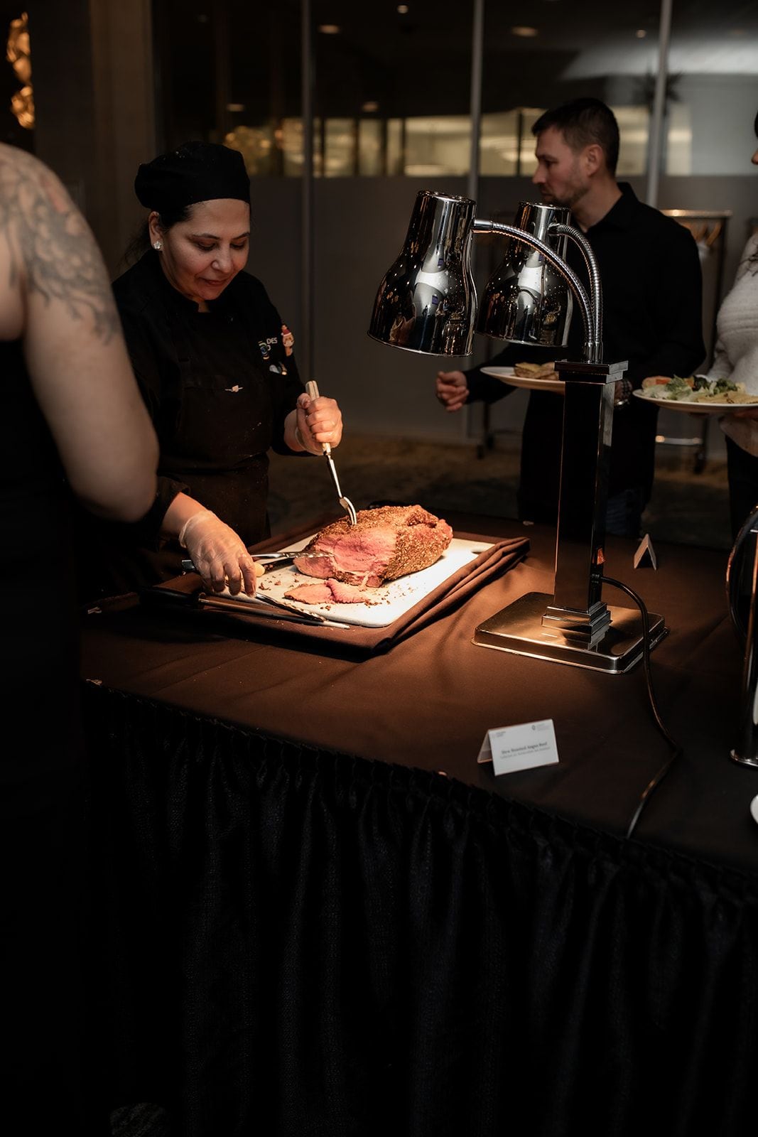 A person carving meat on a cutting board at a table with a lit lamp and a name card.