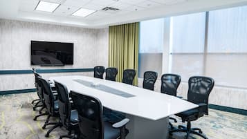 A conference room with a long table, black chairs, a flat-screen TV, and large windows with curtains.