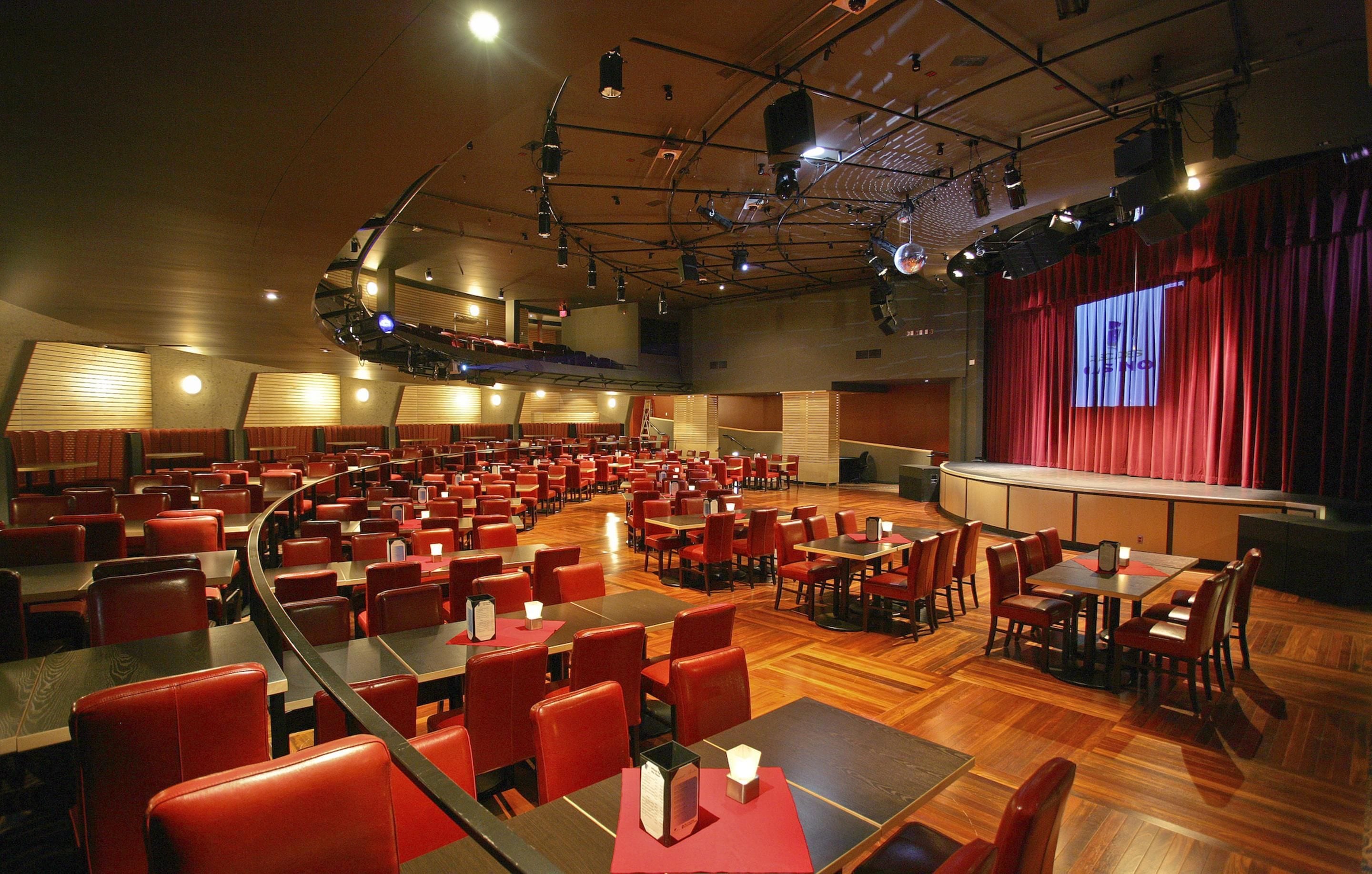 A conference room with multiple tables and chairs, a stage with curtains, and a large screen.