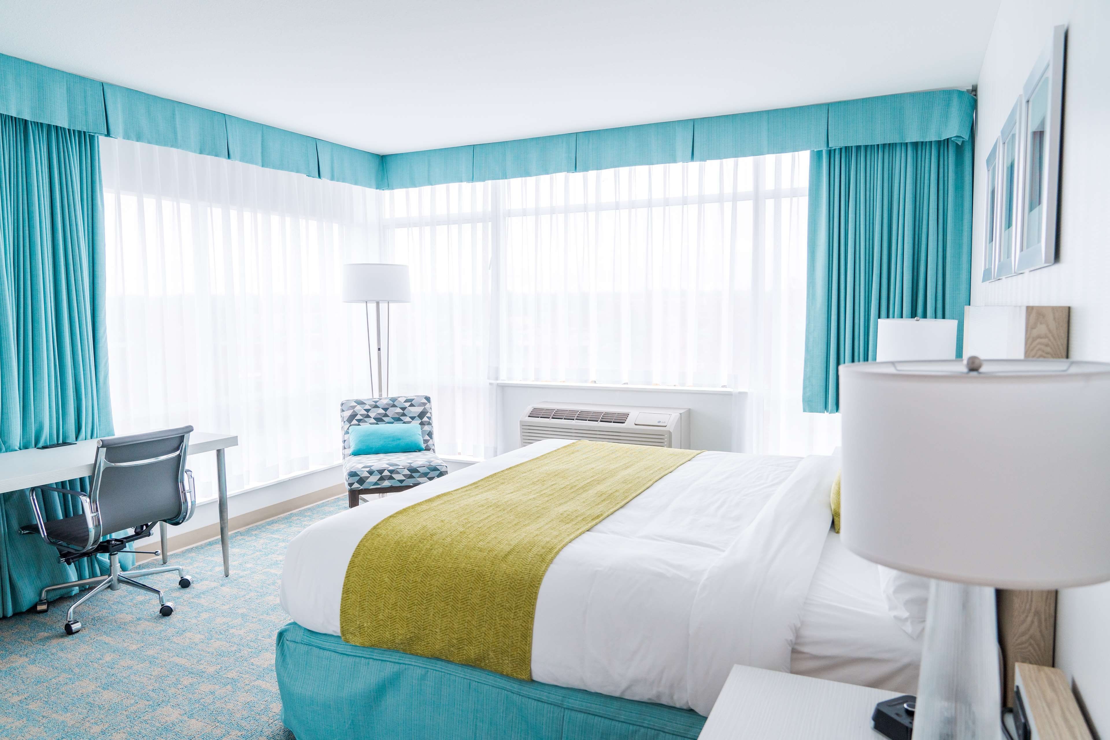 Una habitación de hotel con una cama, un escritorio, una silla, una lámpara y ventanas grandes con cortinas.