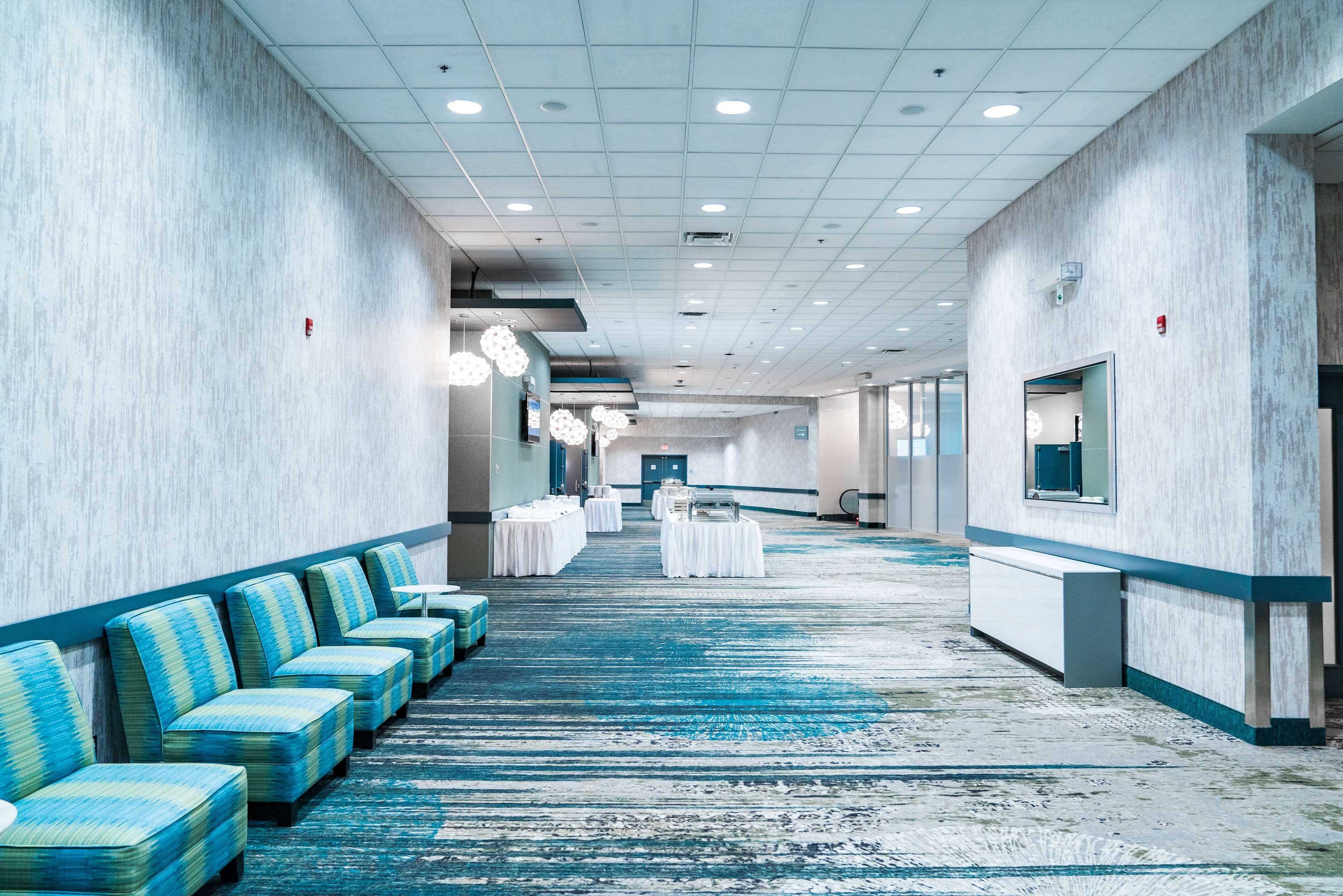 A hotel corridor with rows of blue upholstered seating, a carpeted floor, and a long hallway leading to a reception area.