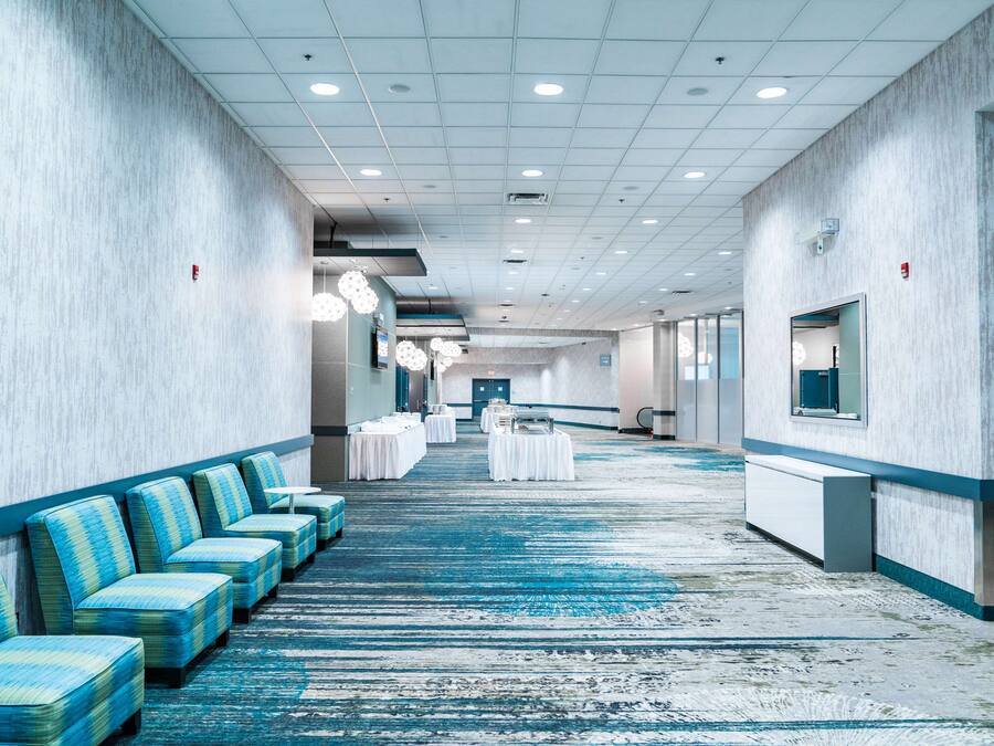 A hotel corridor with rows of blue upholstered seating, a carpeted floor, and a long hallway leading to a reception area.