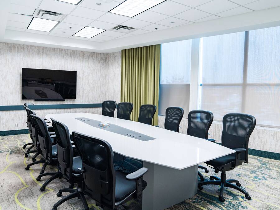 Une salle de conférence avec une longue table, des chaises noires, un téléviseur à écran plat et de grandes fenêtres dotées de rideaux.