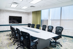 Un salón de conferencias con una larga mesa, sillas negras, un televisor de pantalla plana y ventanas amplias con cortinas.
