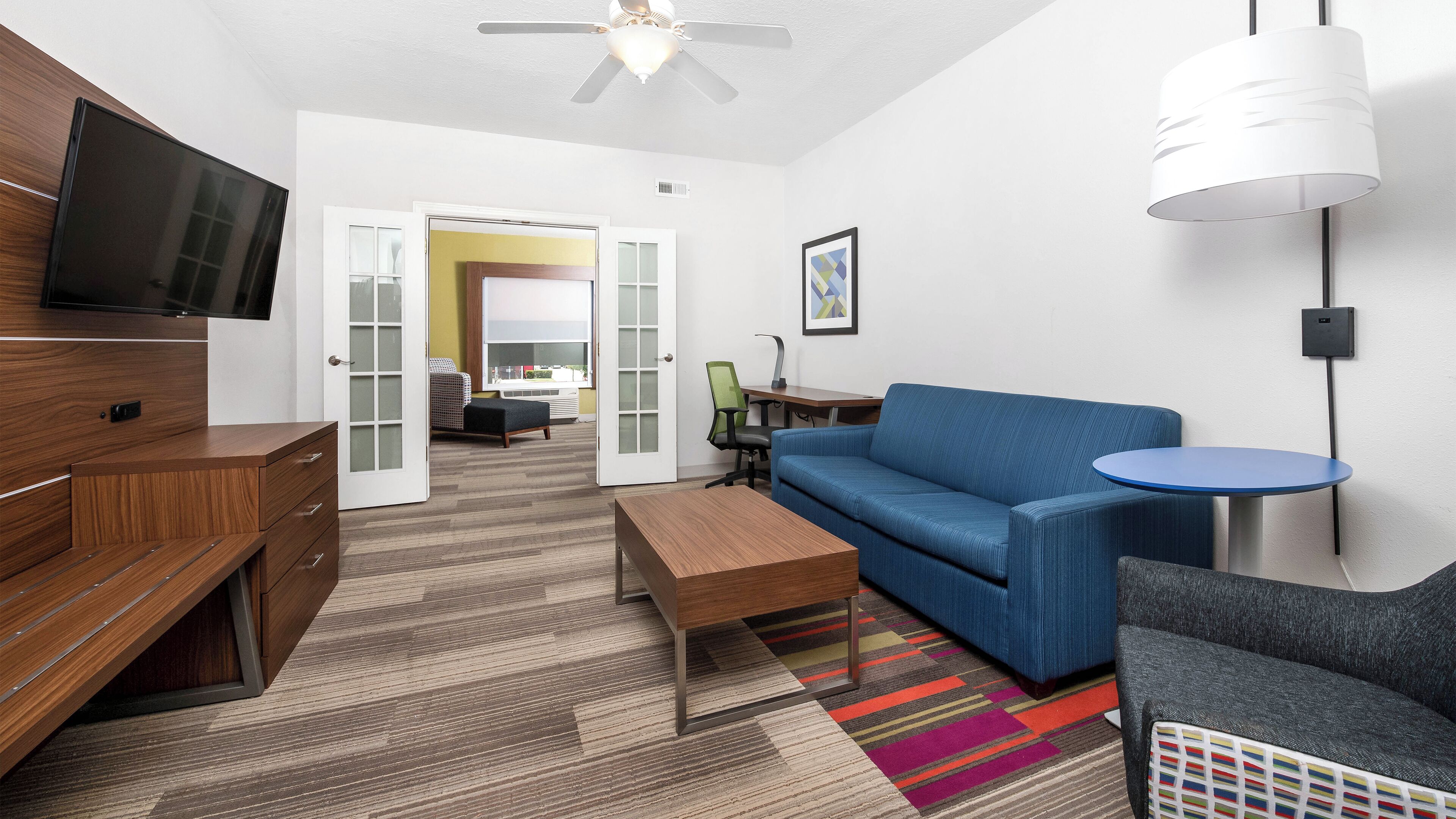 Ein modernes Hotelzimmer mit Flachbildfernseher, einem blauen Sofa, einem kleinen runden Tisch, einer Holzkabine und einem farbenfrohen Teppich.
