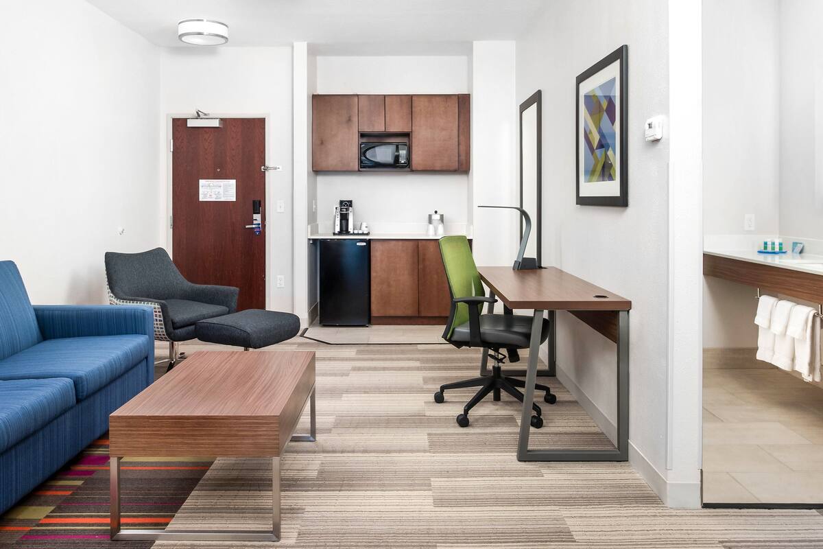 A compact living space with a blue sofa, a wooden coffee table, a green office chair, a kitchenette with a microwave, and a small refrigerator.