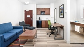A compact living space with a blue sofa, a wooden coffee table, a green office chair, a kitchenette with a microwave, and a small refrigerator. - Holiday Inn Express Hotel & Suites Conroe I-45 North by IHG (Conroe)
