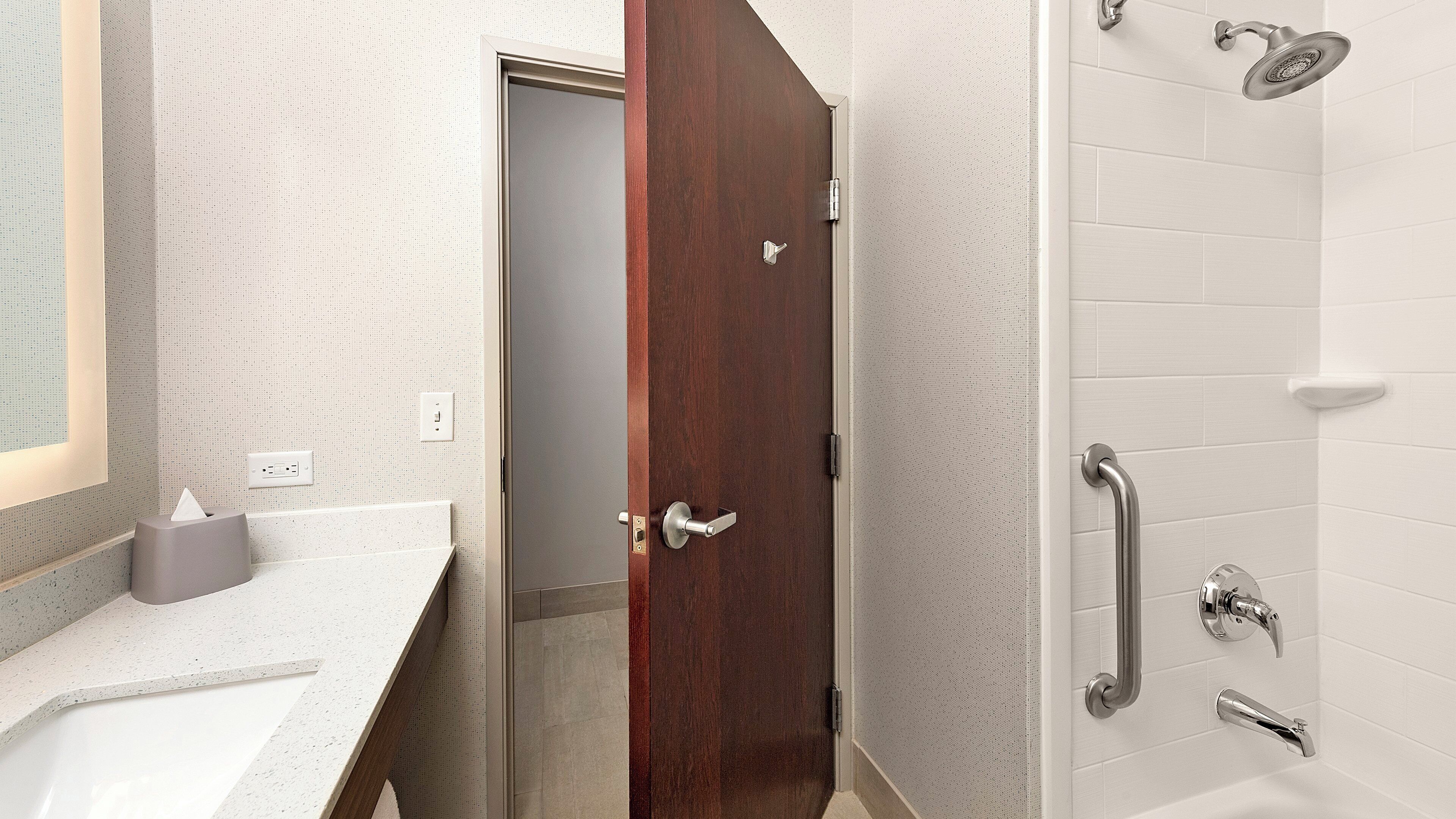 Un baño con puerta de madera, zona de ducha con azulejos blancos, barra de apoyo y toallero.