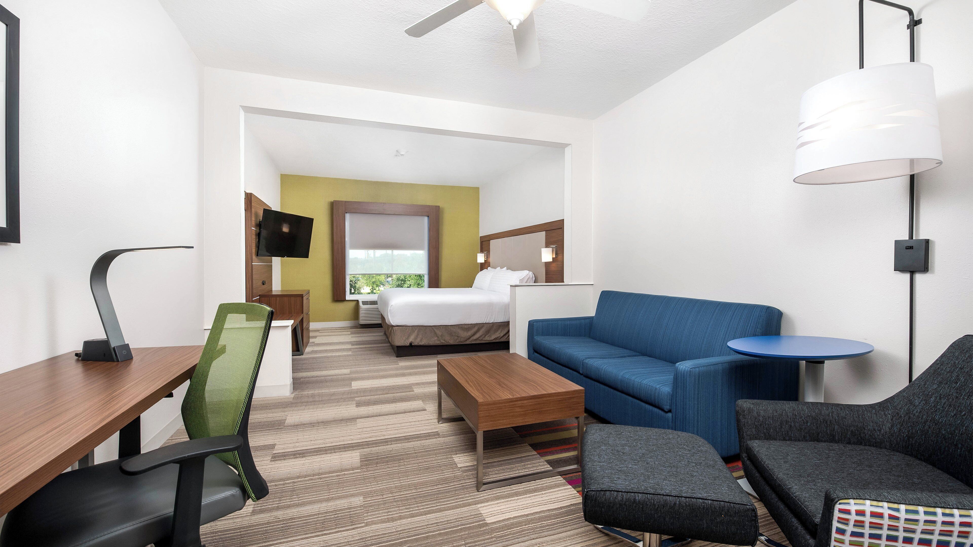 A hotel room with a bed, a sofa, a desk, a chair, a TV, and a lamp.