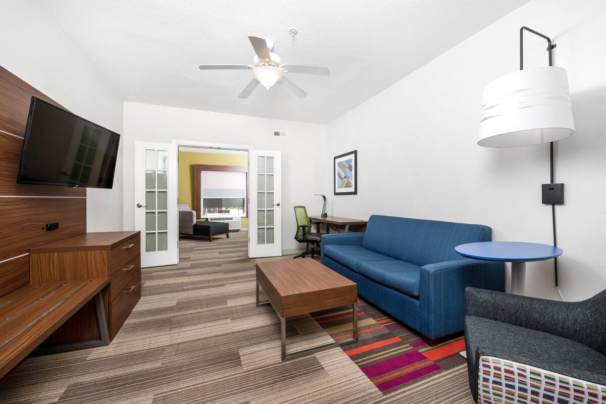 A modern hotel room with a flat-screen TV, a ceiling fan, a sofa, a coffee table, a desk, and a chair.
