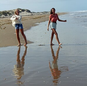 Dos personas caminando sobre una playa arenosa, con el agua reflejando sus imágenes.