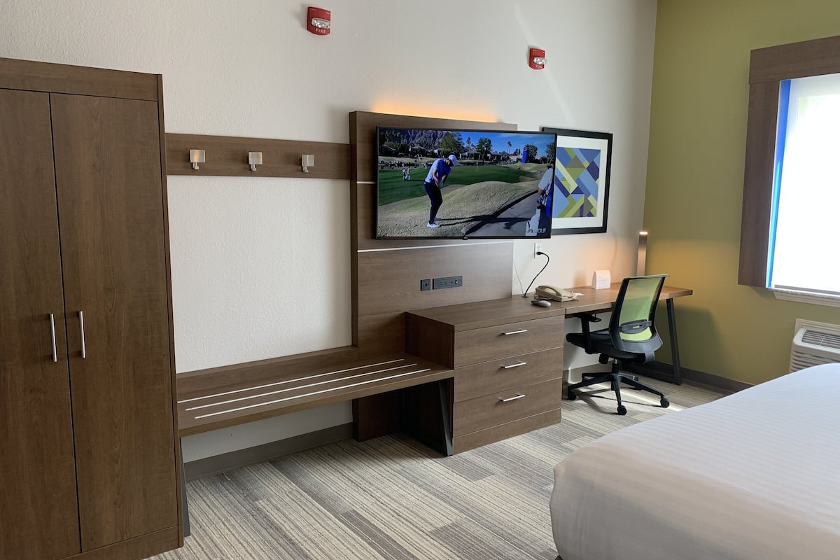 A hotel room with a bed, a desk with a computer, a television mounted on the wall, and a wardrobe.