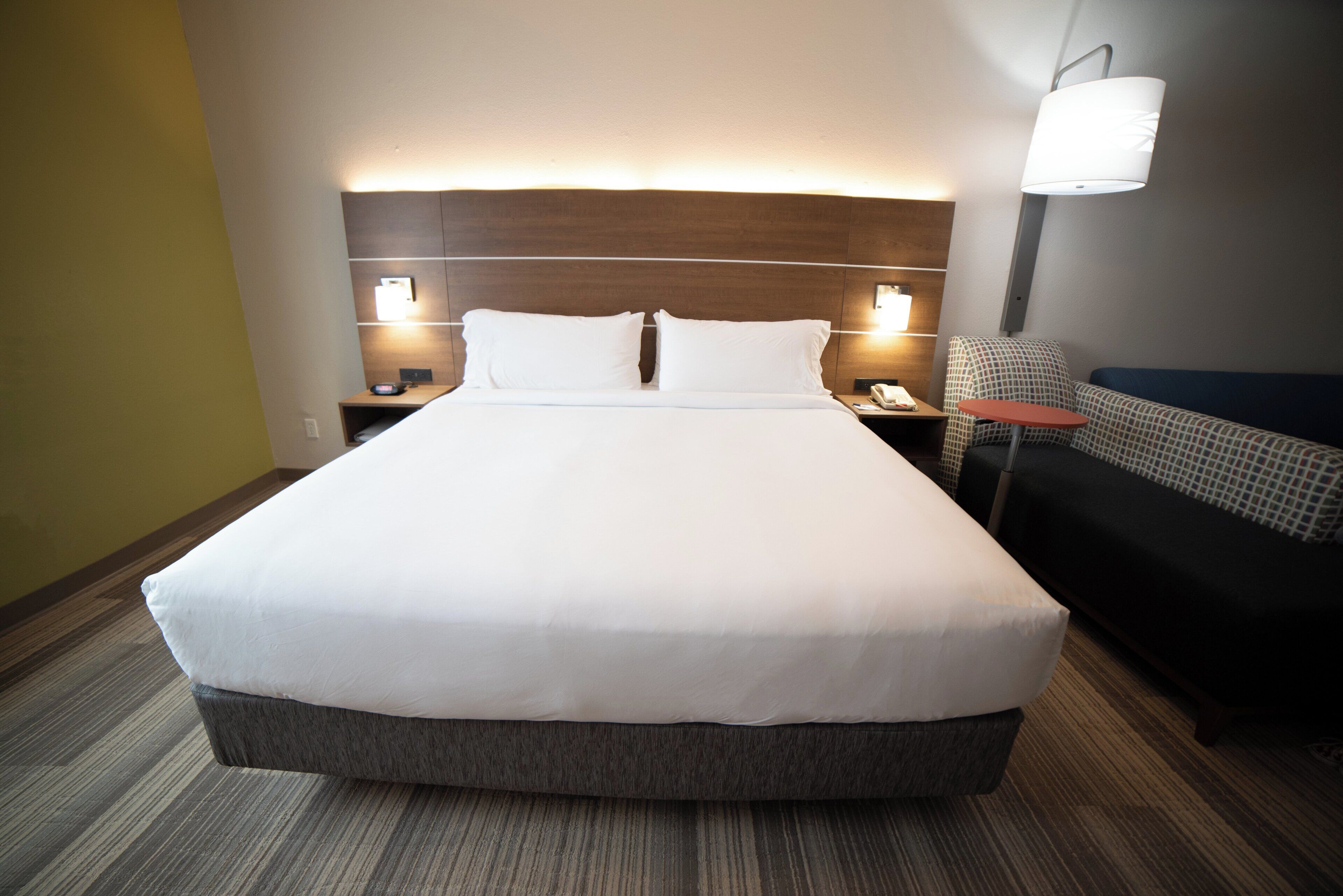 Una cama grande con cabecera de madera, dos lámparas de noche, una mesita y un sofá en una habitación de hotel.