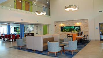 A spacious lobby with a high ceiling, a seating area with patterned sofas, a dining area with red chairs, and a large aquarium on the wall.