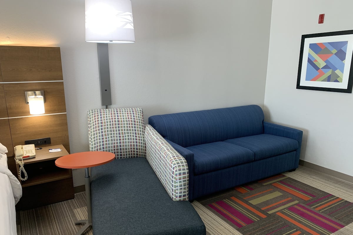 A compact hotel room with a blue sofa, a small round table, a checkered armchair, a patterned rug, and a wall-mounted lamp.