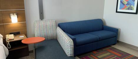 Ein Hotelzimmer mit einem blauen Sofa, einem kleinen Tisch, einem karierten Sessel, einem gemusterten Teppich und einer Wandlampe.