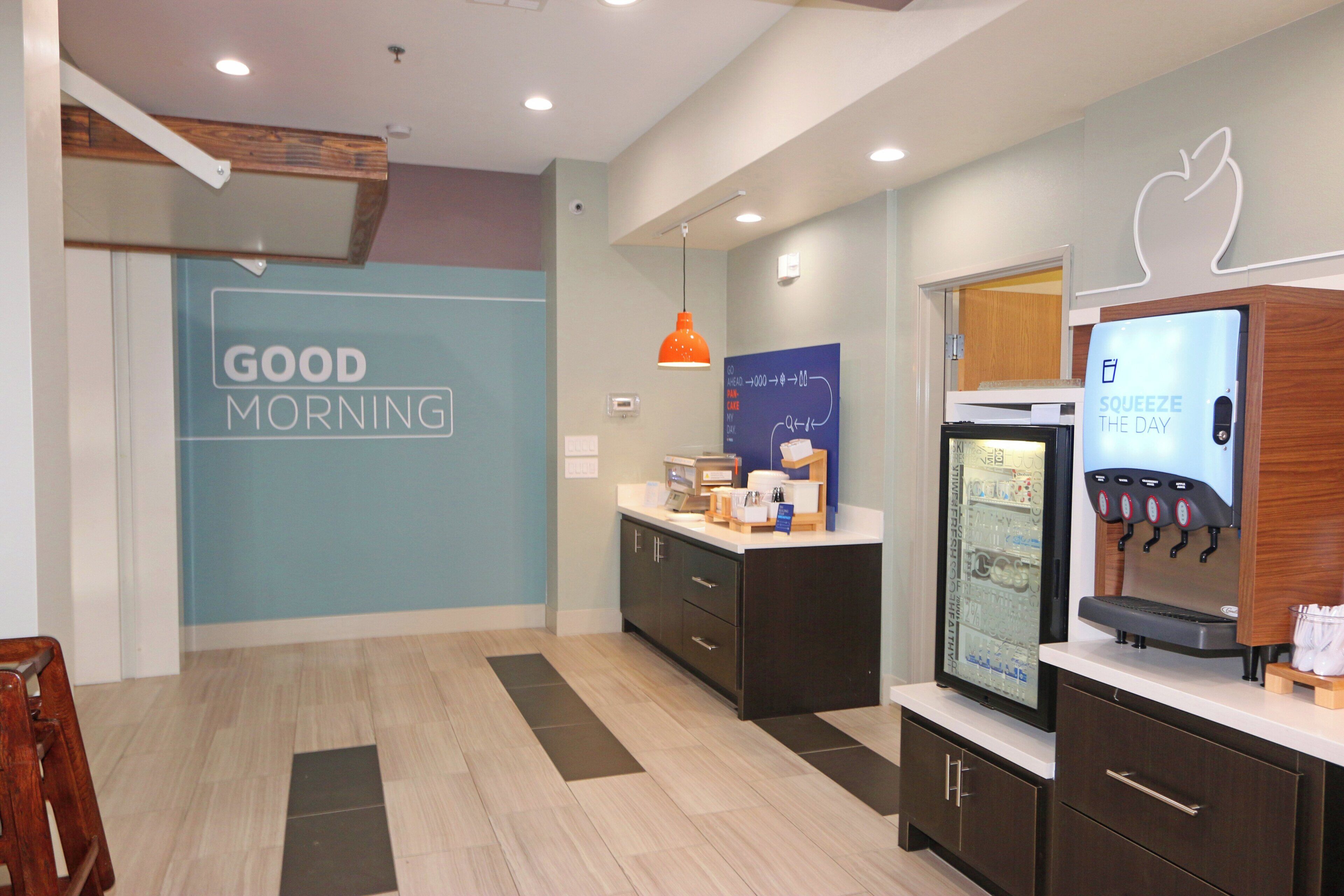 A modern kitchen area with a 'GOOD MORNING' sign, a coffee machine, and a refrigerator with a branded display.