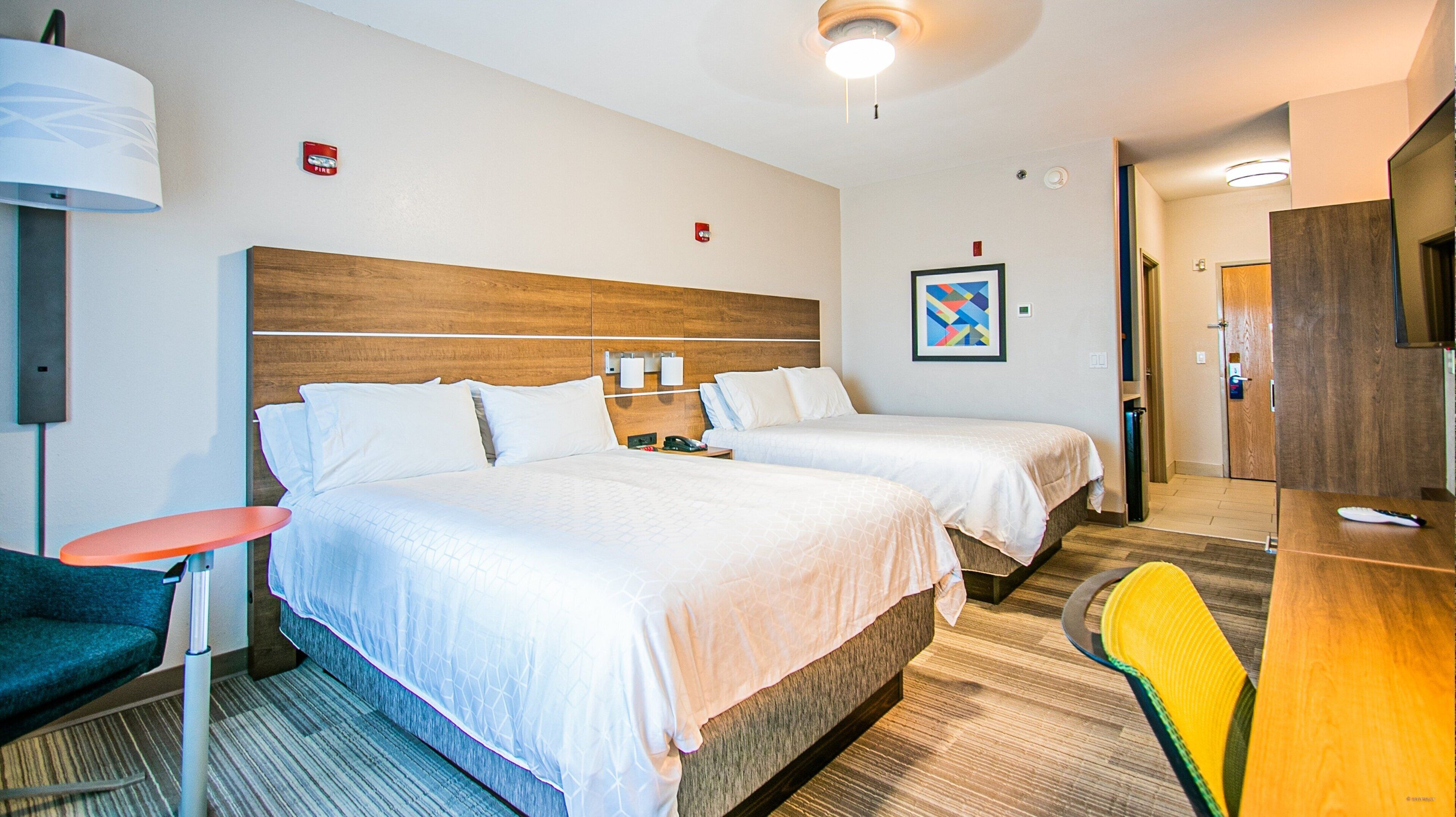 A hotel room with two beds, a desk, a chair, a lamp, and a fire alarm on the wall.