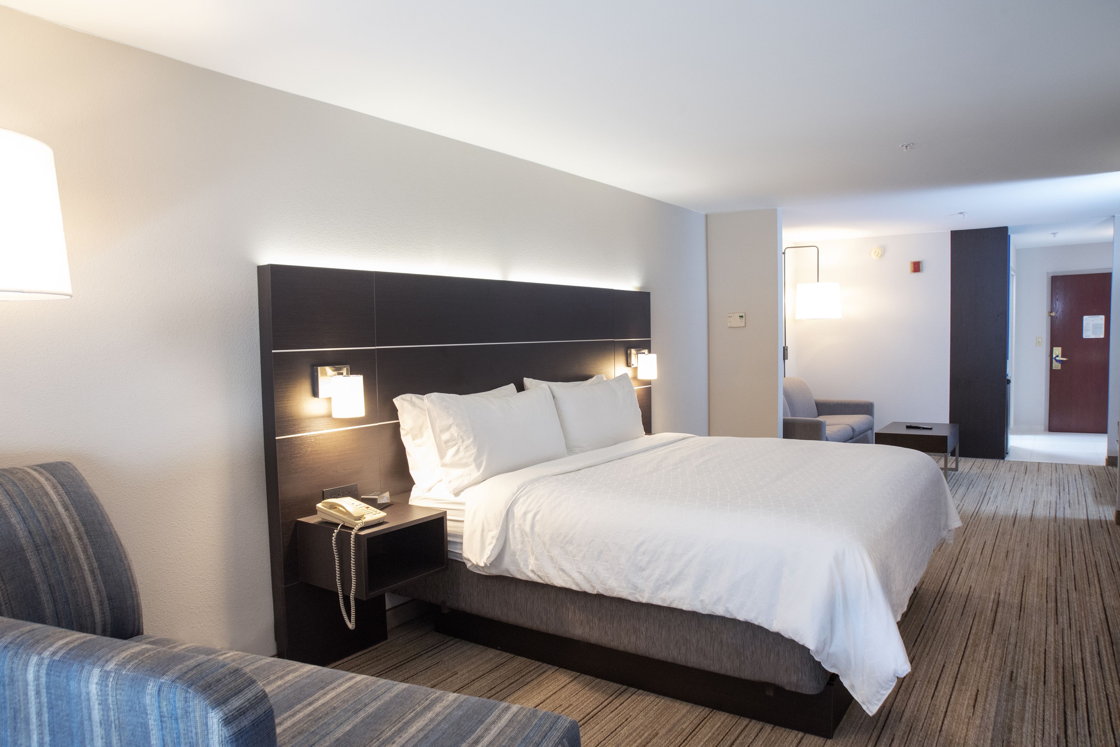 A modern hotel room with a large bed, a bedside table with a lamp, a chair, and a desk with a phone.