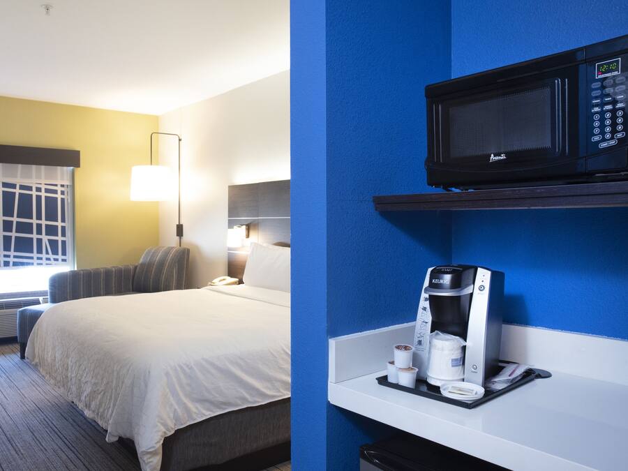 Une chambre d’hôtel comprenant un lit, une table de chevet, une lampe, une fenêtre avec stores, un four à micro-ondes fixé au mur et une machine à café posée sur un plateau.