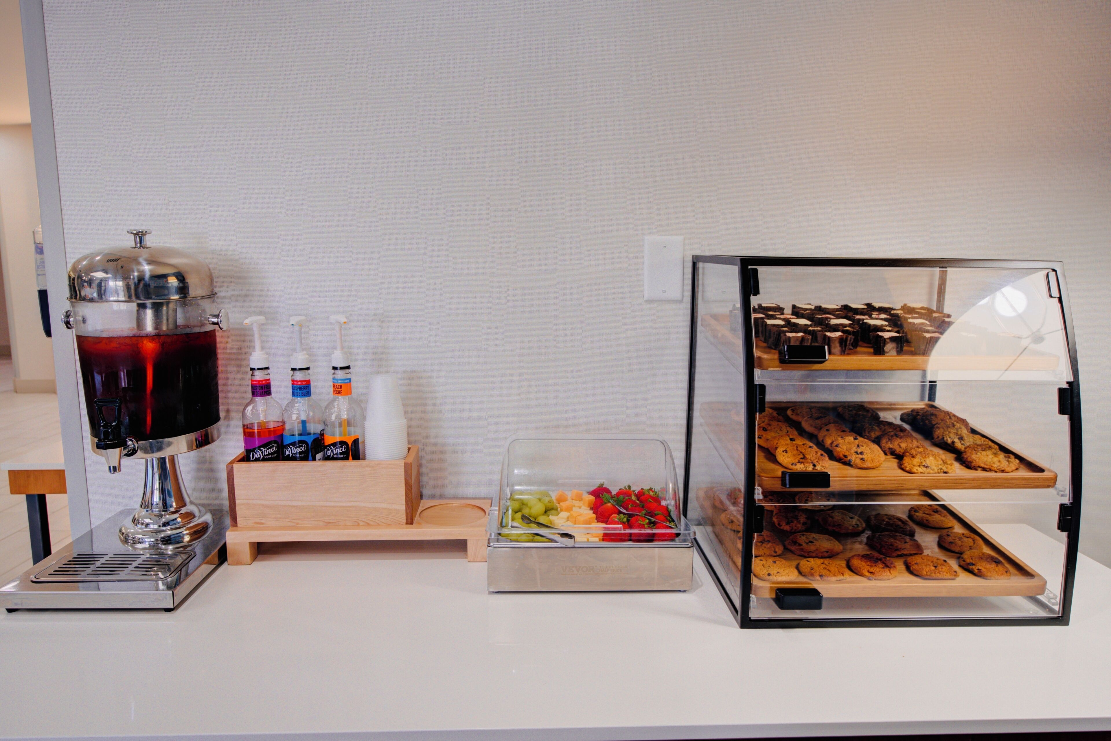 En buffé med en kaffemaskin, en dryckesdispenser och ett varierat utbud av matvaror, inklusive frukt, grönsaker och bakverk.