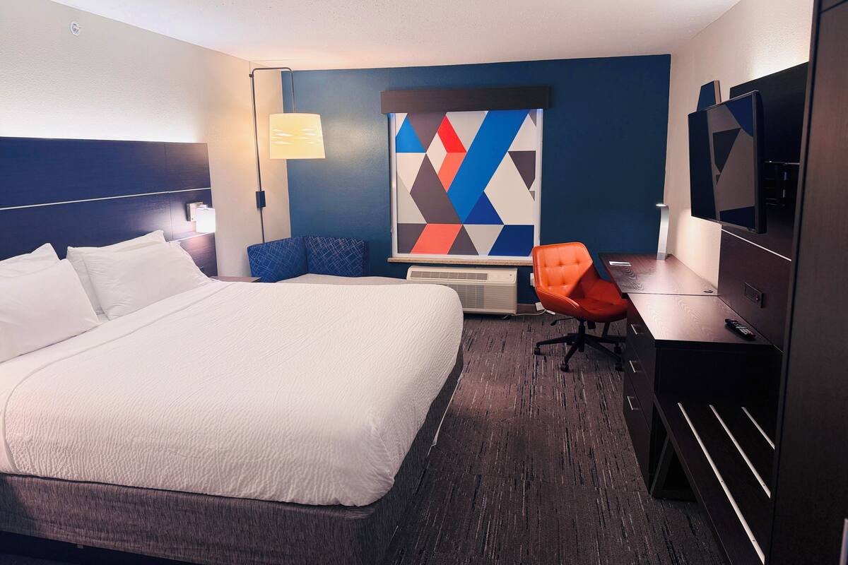 A hotel room with a large bed, a desk with a chair, a TV, and a wall-mounted air conditioner.