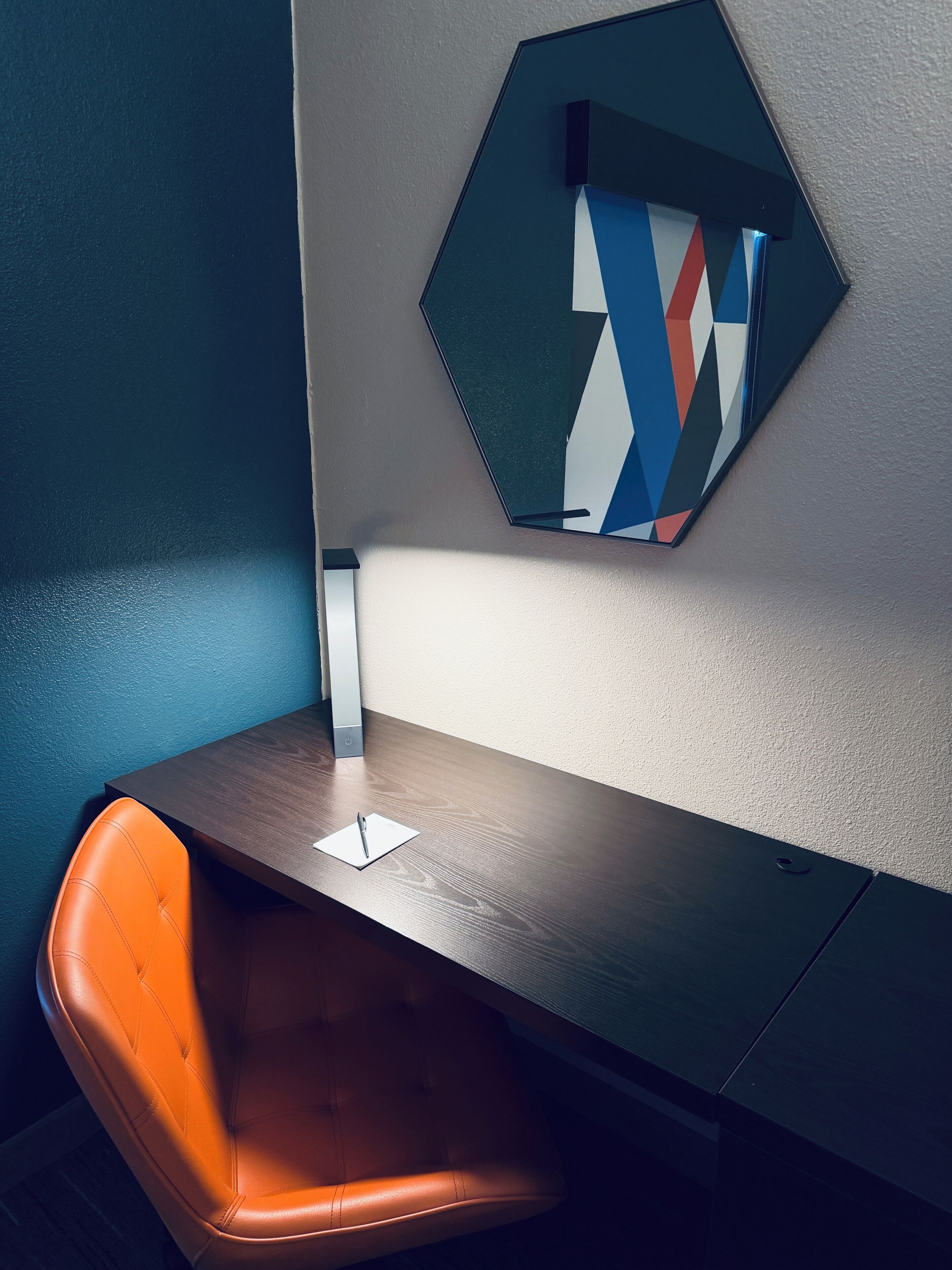 A corner of a room with a wooden table, a single orange chair, and a geometric-patterned wall decoration.
