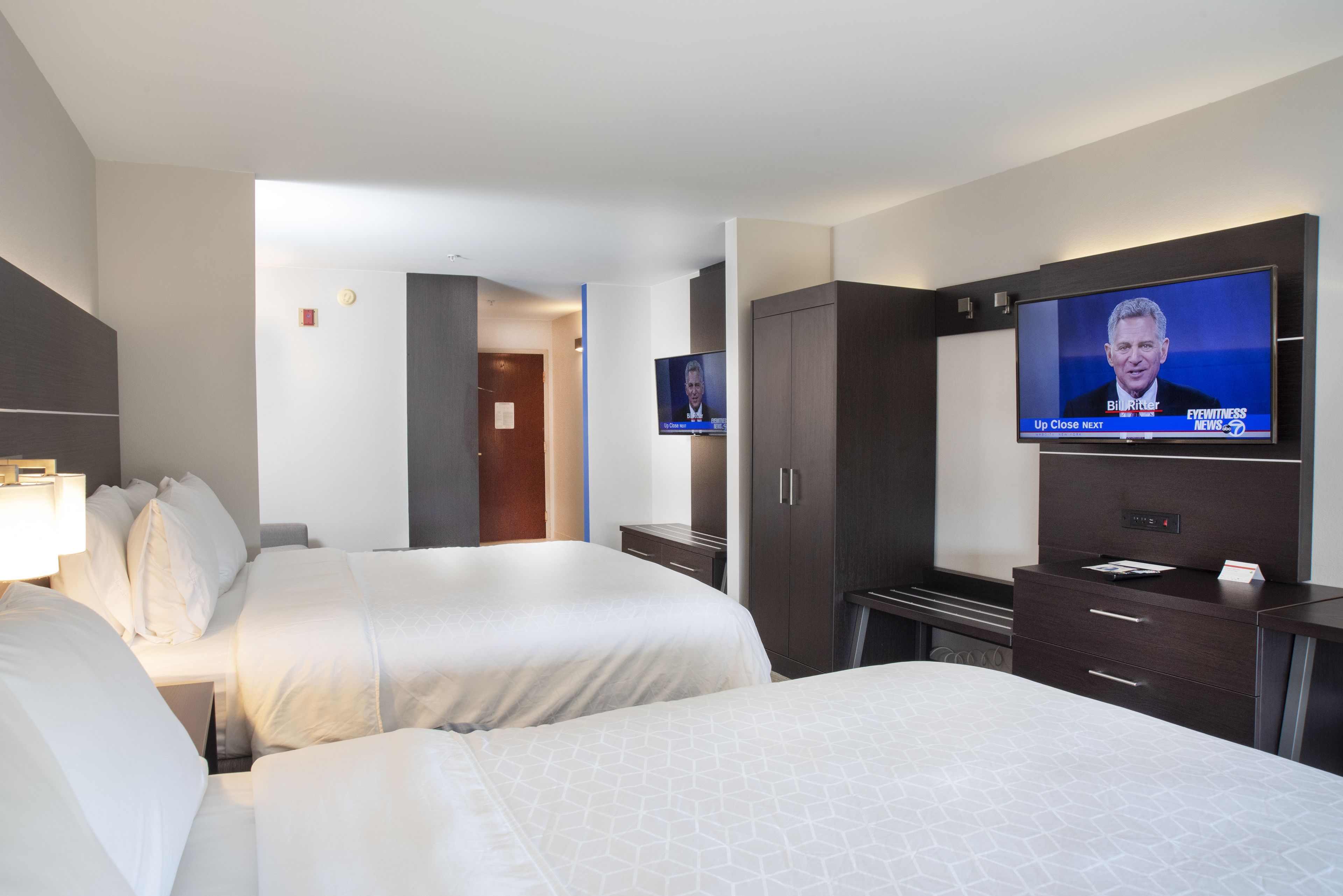 Habitación de hotel con dos camas, un televisor mostrando un noticiero y un armario.