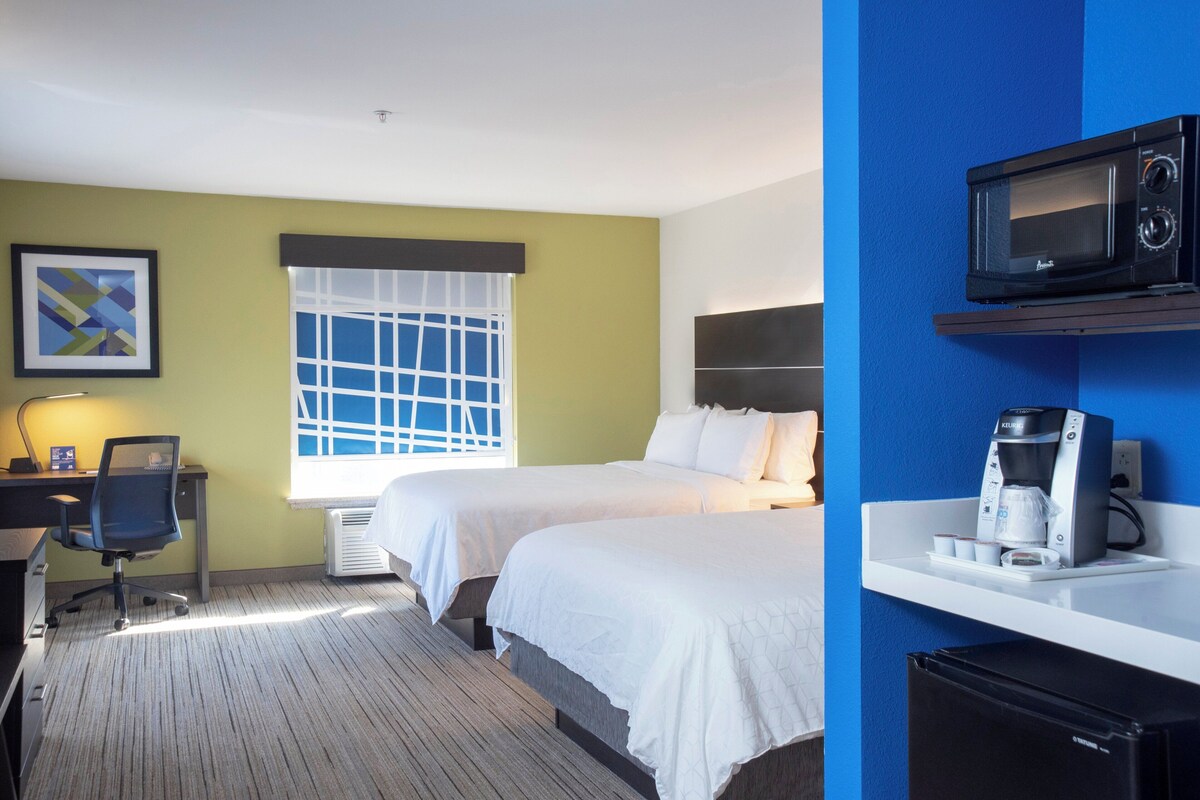 Une chambre d’hôtel avec deux lits, un bureau avec une chaise, une fenêtre avec des stores, un four à micro-ondes et une machine à café.