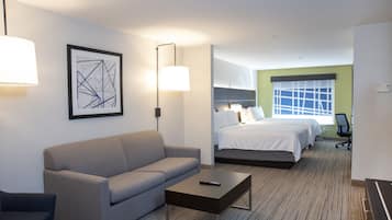 A hotel room with a grey sofa, a small table, a bed with white bedding, a desk with a chair, and a framed abstract artwork on the wall.