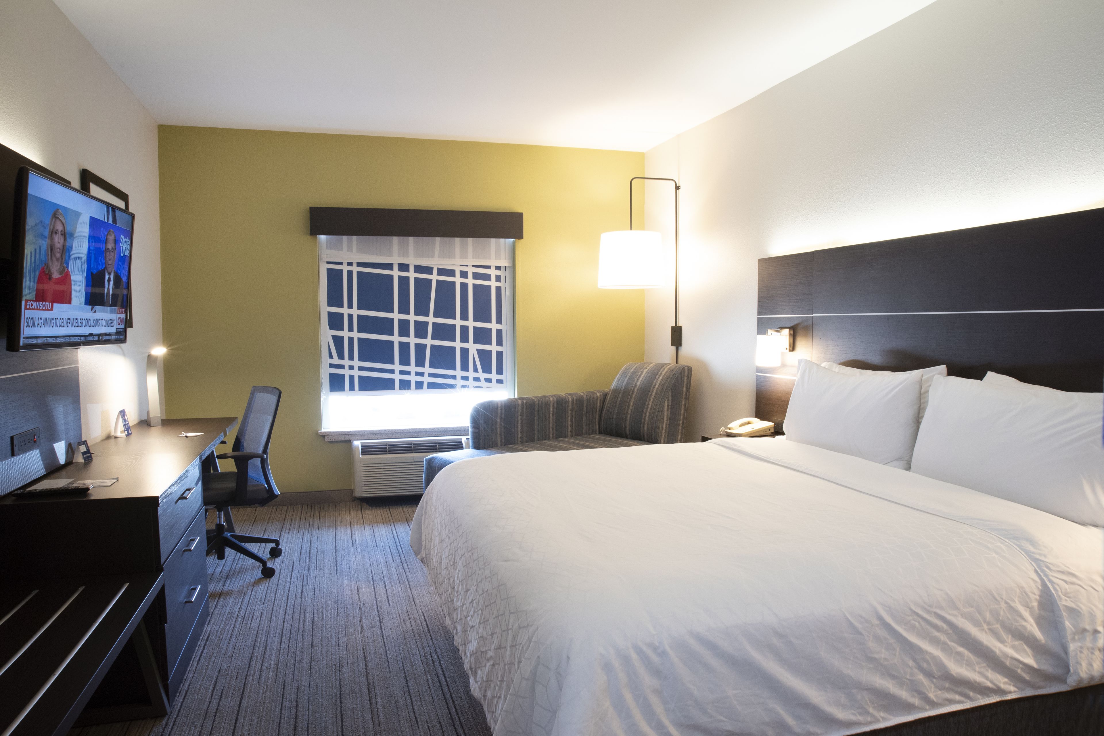 A hotel room with a large bed, a desk with a computer, a chair, a TV displaying news, and a window with blinds.
