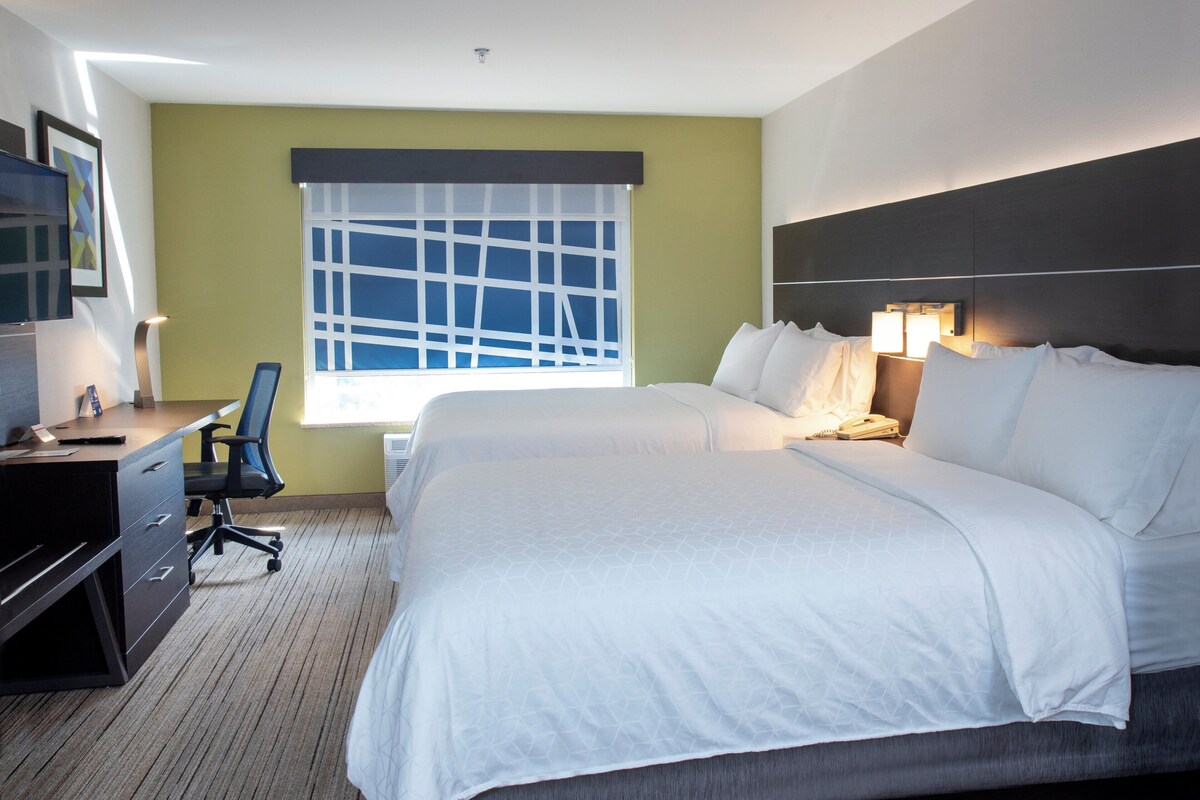 Une chambre d’hôtel avec un grand lit, un bureau avec une chaise, une fenêtre avec des stores et une tête de lit dotée de lampes.