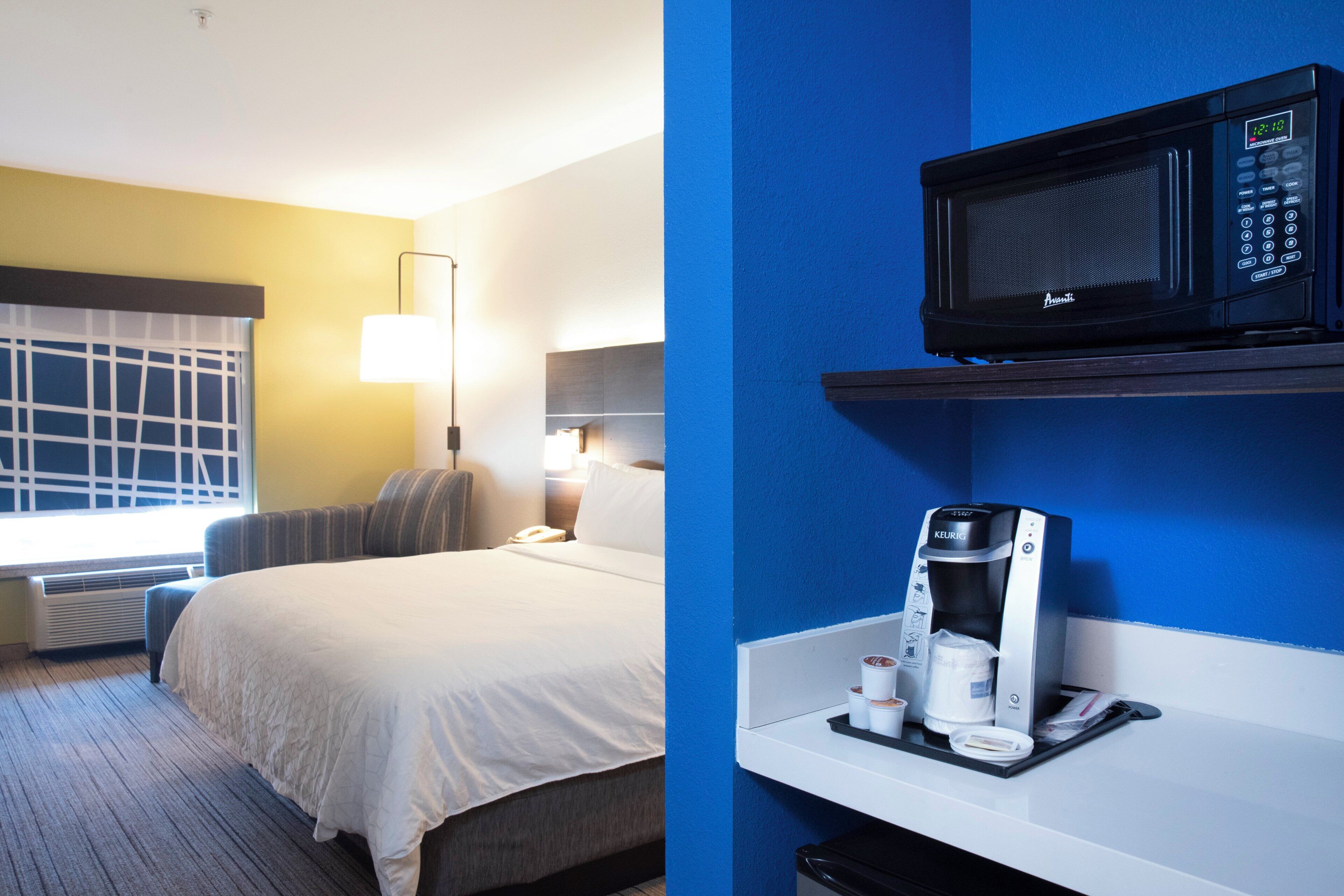 Habitación de hotel con cama, lámpara de noche, ventana y una kitchenette con microondas y cafetera.