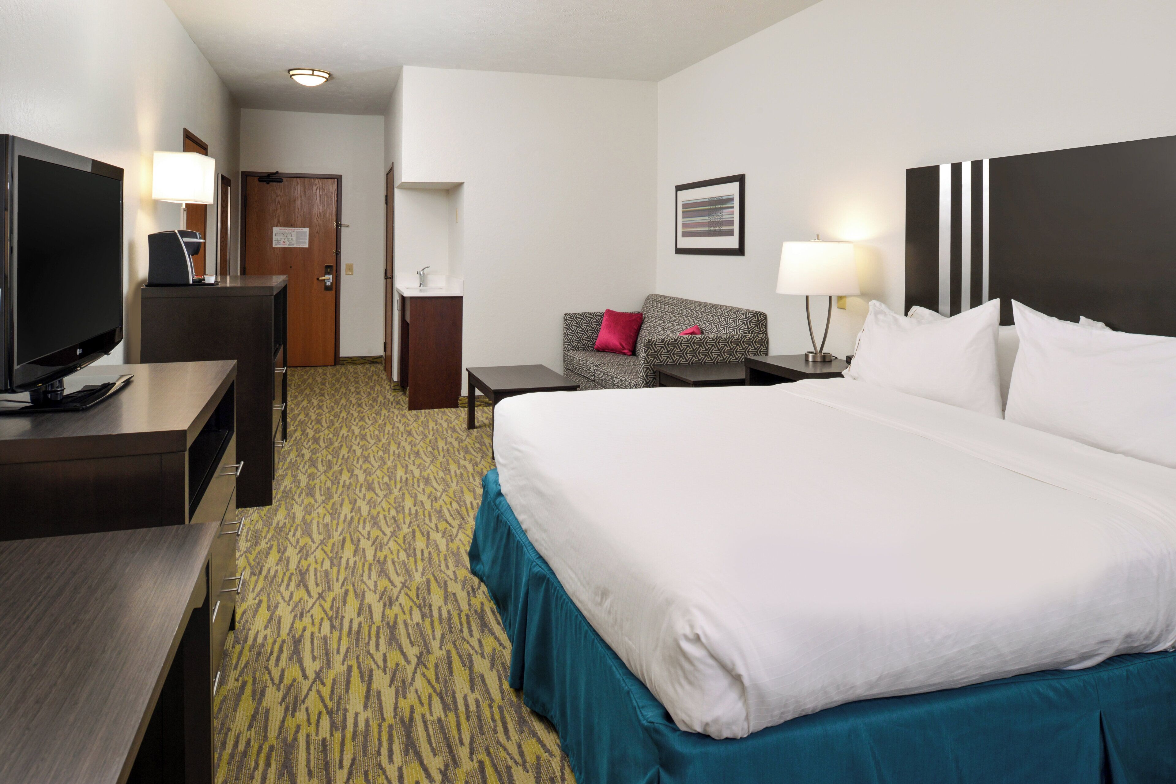 A hotel room with a large bed, a desk with a flat-screen TV, a sofa, a small table, and a wall-mounted picture.