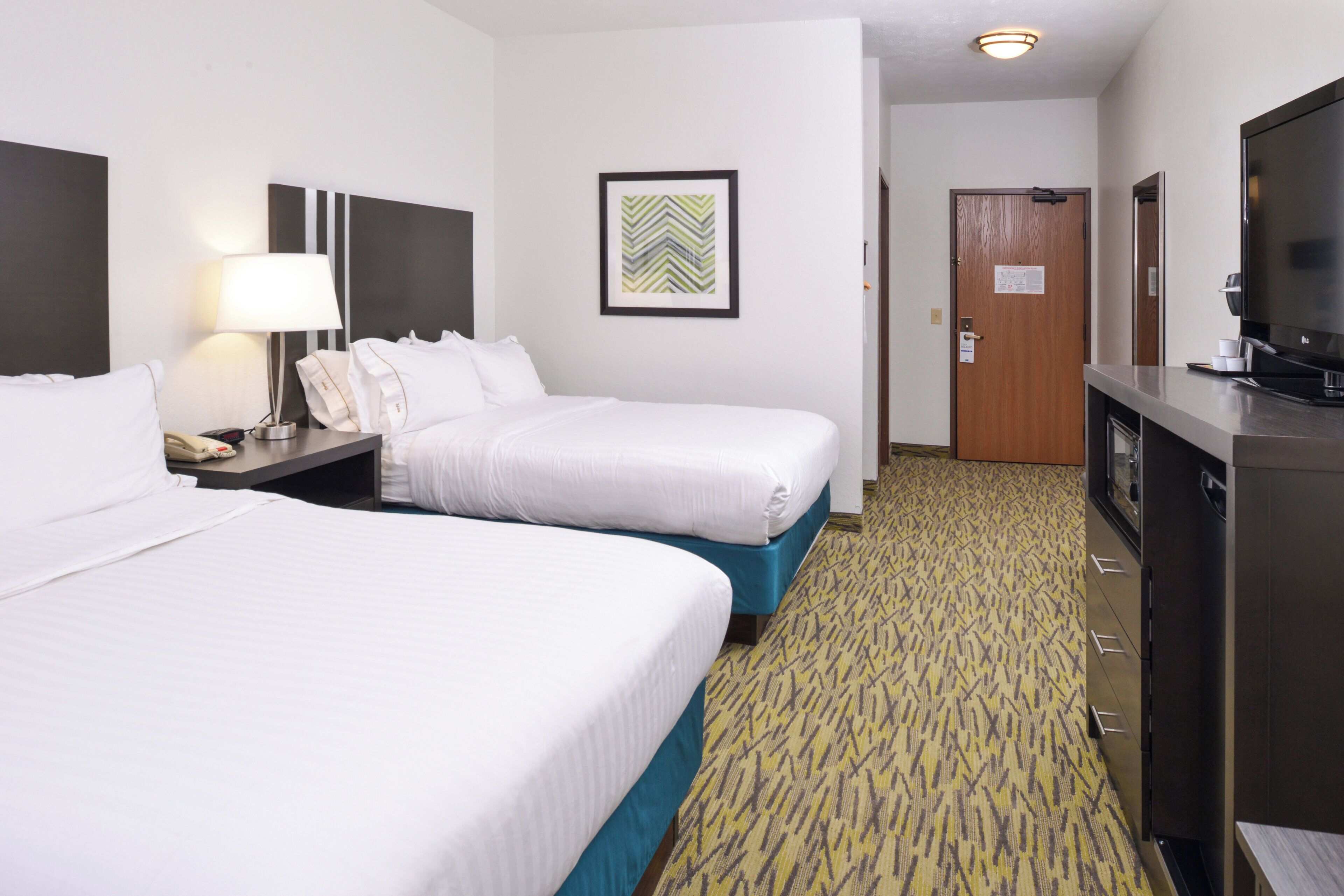 A hotel room with two beds, a desk with a lamp, a television, and a door.
