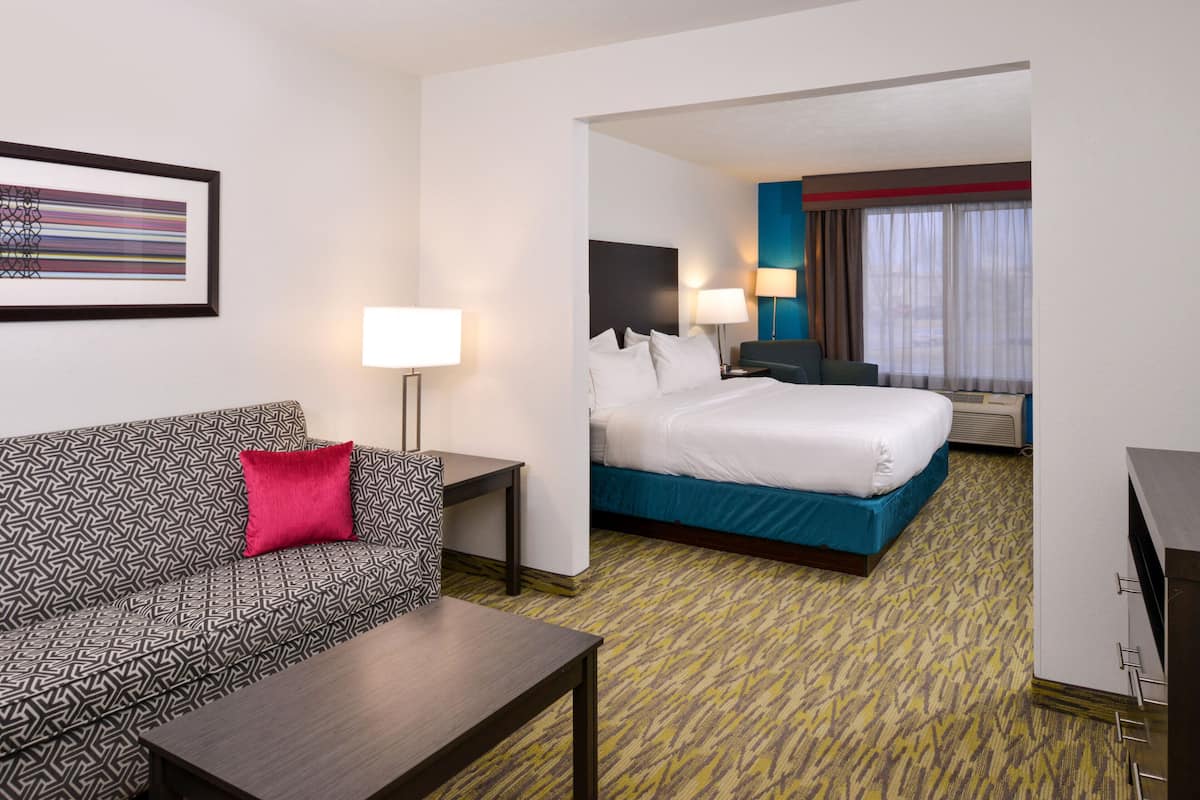 A hotel room with a patterned sofa, a bed with a blue headboard, a nightstand, a lamp, and a framed picture on the wall.