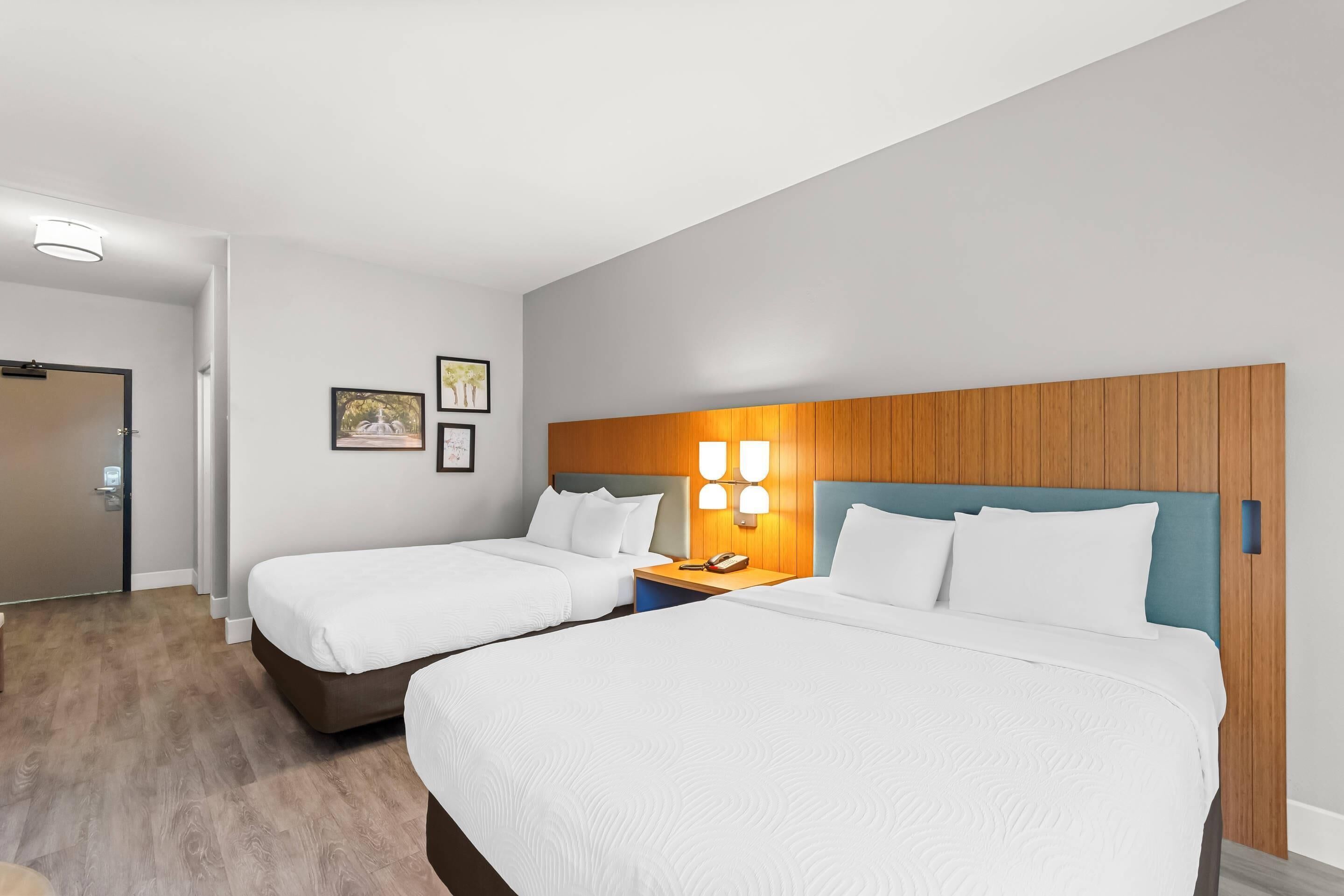 A hotel room with two beds, a wooden headboard, a nightstand with a lamp, and framed pictures on the wall.