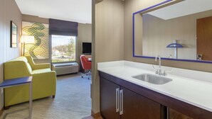 Una habitación de hotel moderna con un ventanal grande, un sofá, un escritorio y un baño con lavamanos y espejo.