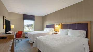 A hotel room with two beds, a desk with a TV, a red chair, and a window with a view of buildings.