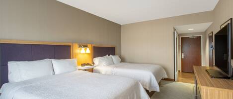 A hotel room with two beds, a desk with a flat-screen TV, a bedside table with a lamp, and a door leading to another room.
